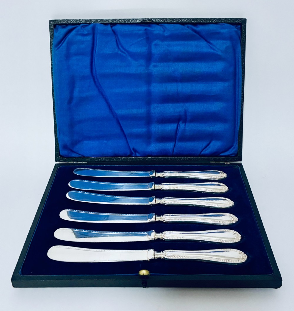 A cased set of six silver-handled butter knives with rounded blades displayed in a dark velvet-lined presentation box.