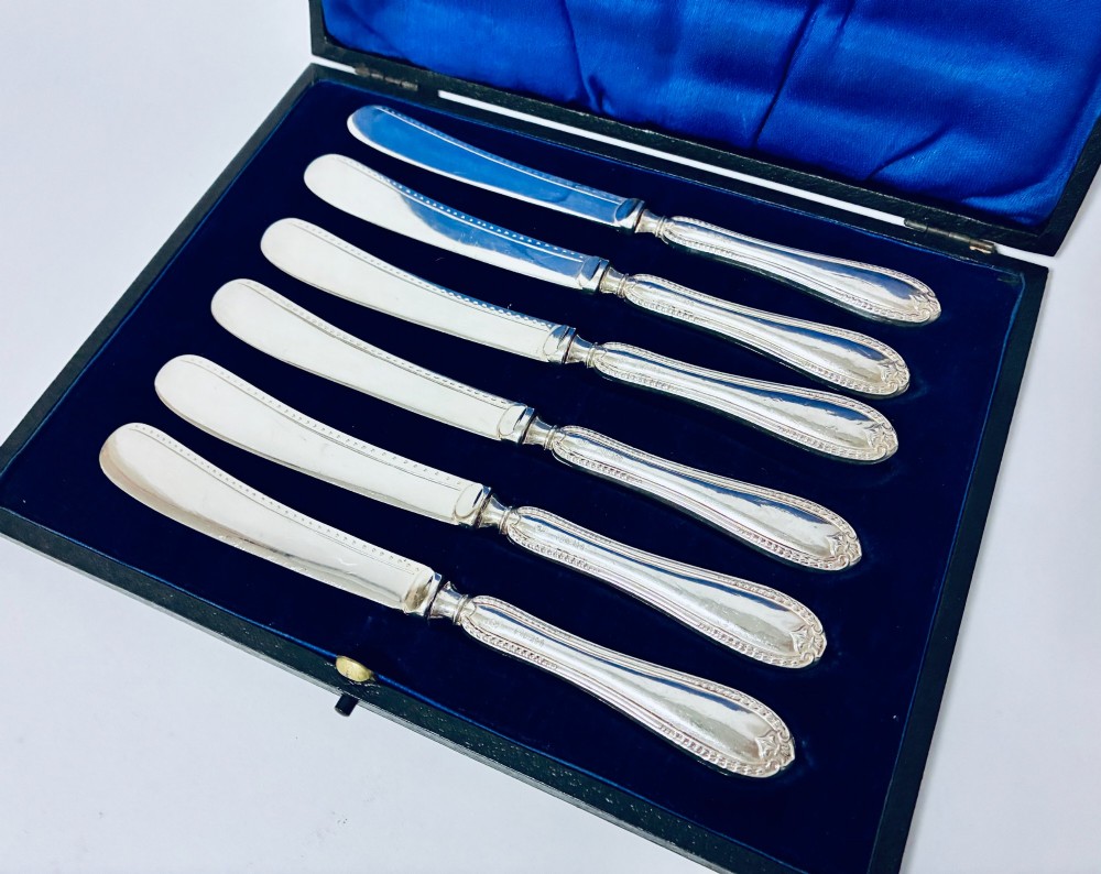 A cased set of six silver-handled butter knives with rounded blades displayed in a dark velvet-lined presentation box. View II