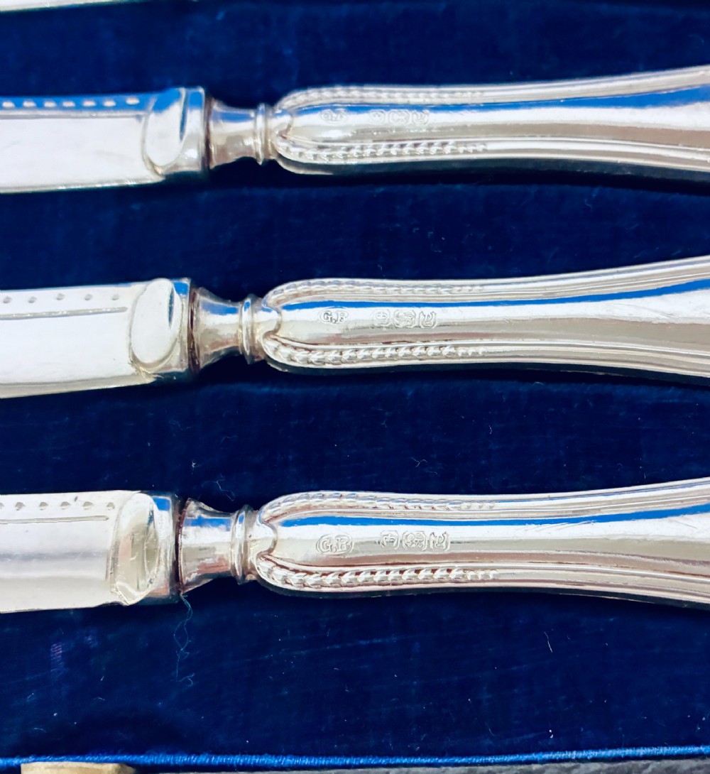 A cased set of six silver-handled butter knives with rounded blades displayed in a dark velvet-lined presentation box. View V