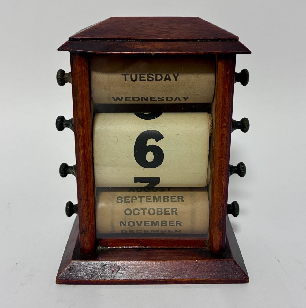 An antique wooden perpetual desk calendar featuring rotating printed scrolls for days, dates, and months displayed behind glass with turning knobs.