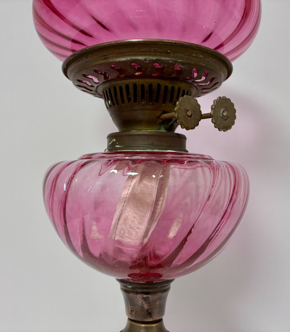 A Victorian oil lamp featuring a swirl optique cranberry glass shade and reservoir set atop a brass column and relief-moulded square base. View II