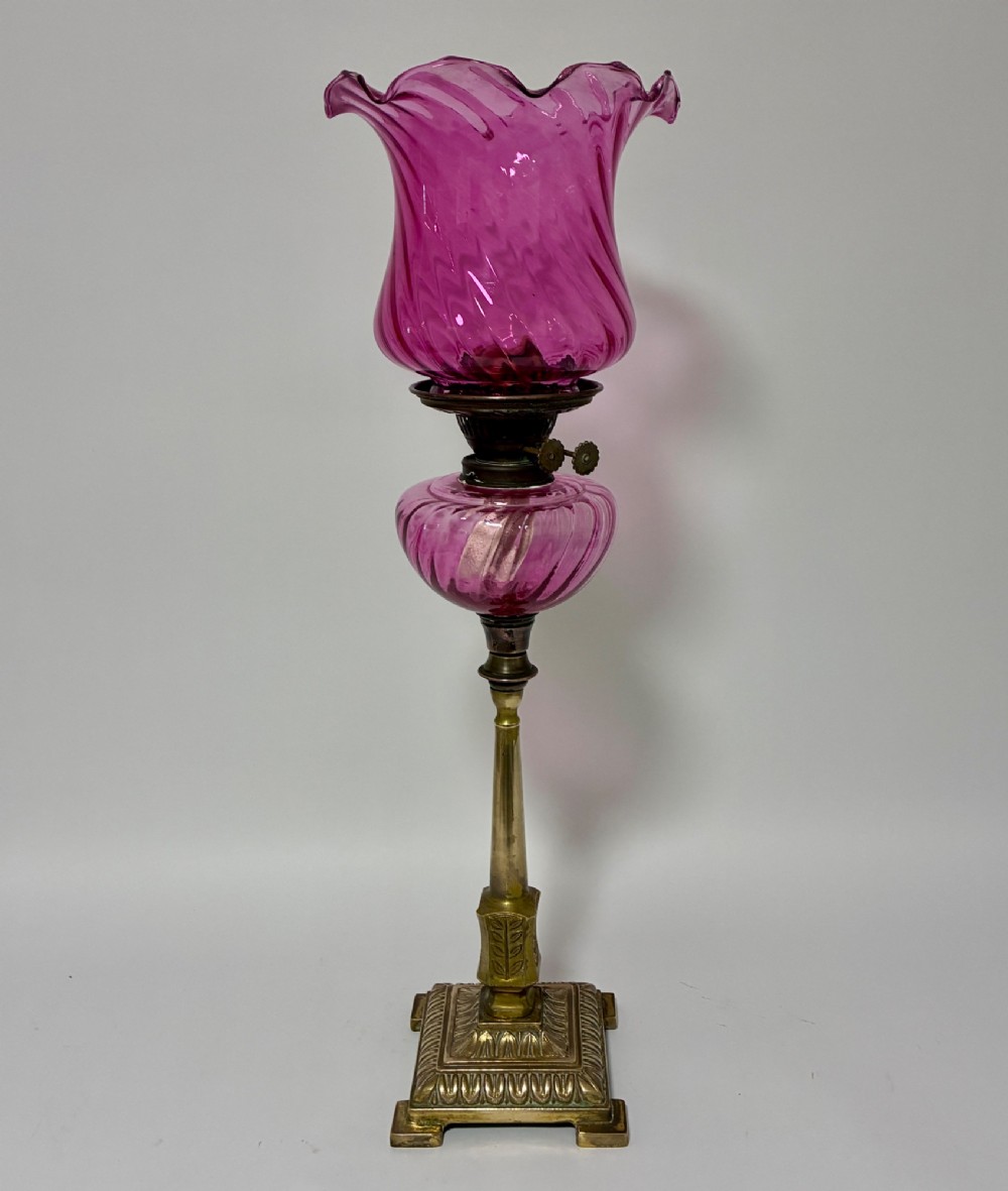 A Victorian oil lamp featuring a swirl optique cranberry glass shade and reservoir set atop a brass column and relief-moulded square base.