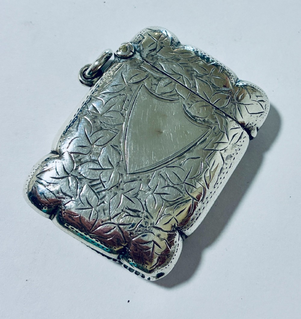 A Victorian sterling silver vesta case featuring hand-engraved foliage motifs, a central shield cartouche, lobed edges, and a side attachment ring.