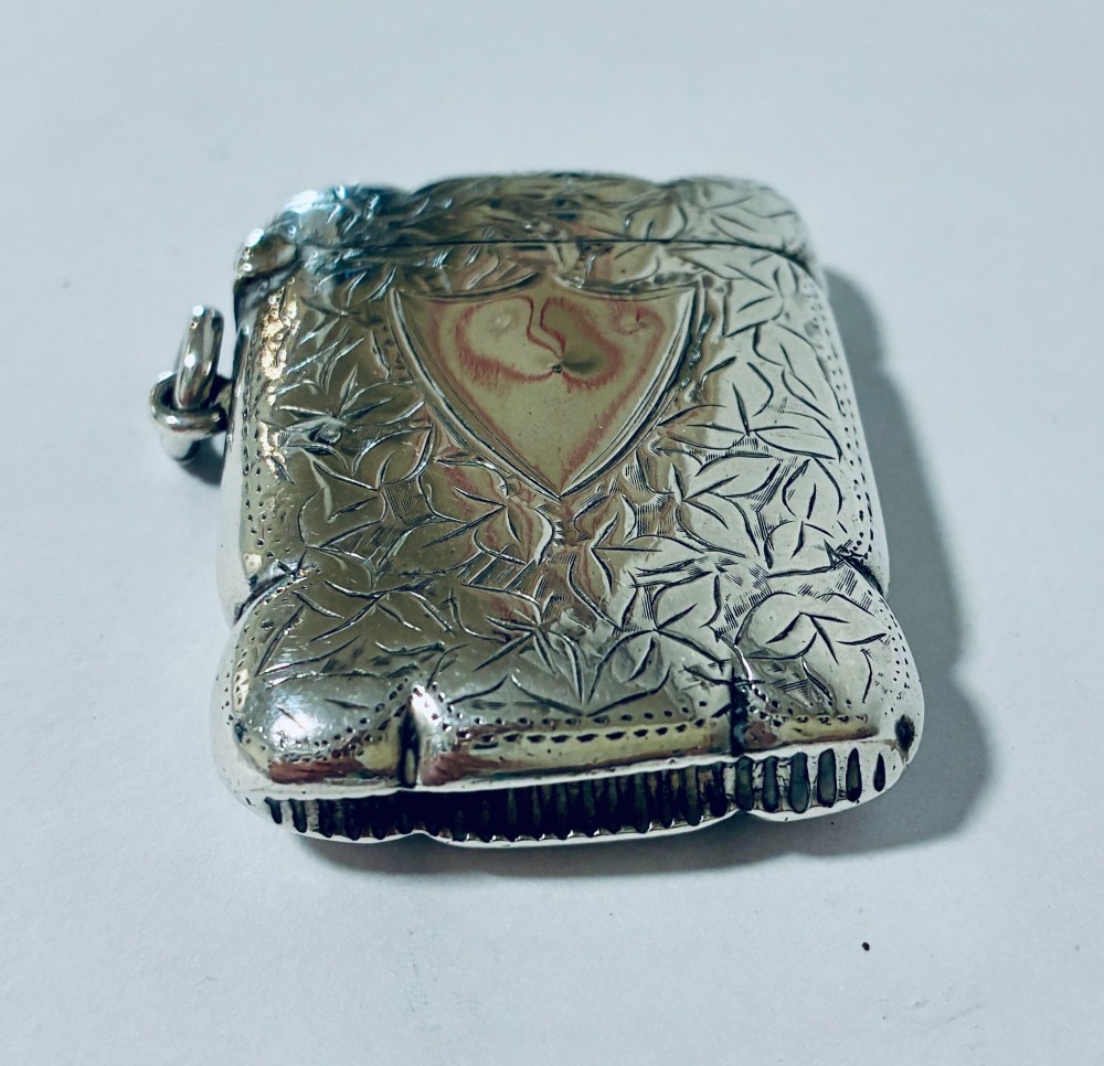 A Victorian sterling silver vesta case featuring hand-engraved foliage motifs, a central shield cartouche, lobed edges, and a side attachment ring. View II