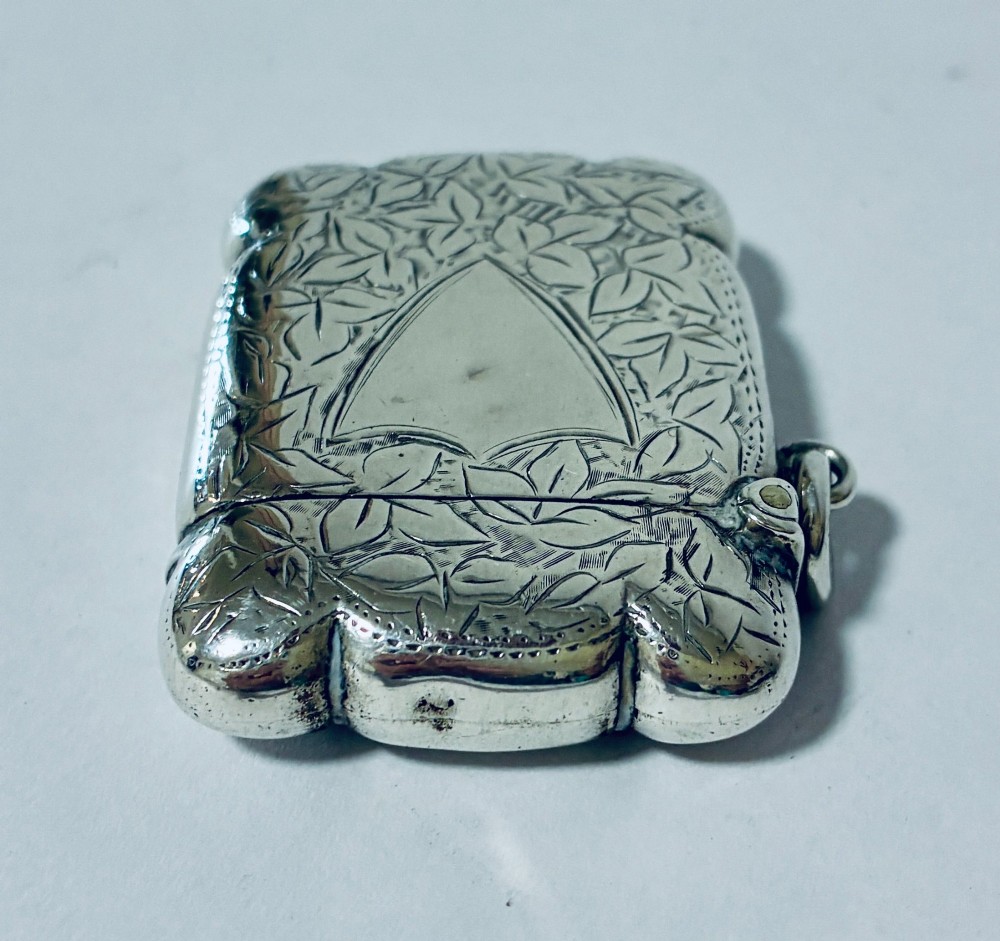 A Victorian sterling silver vesta case featuring hand-engraved foliage motifs, a central shield cartouche, lobed edges, and a side attachment ring. View IV