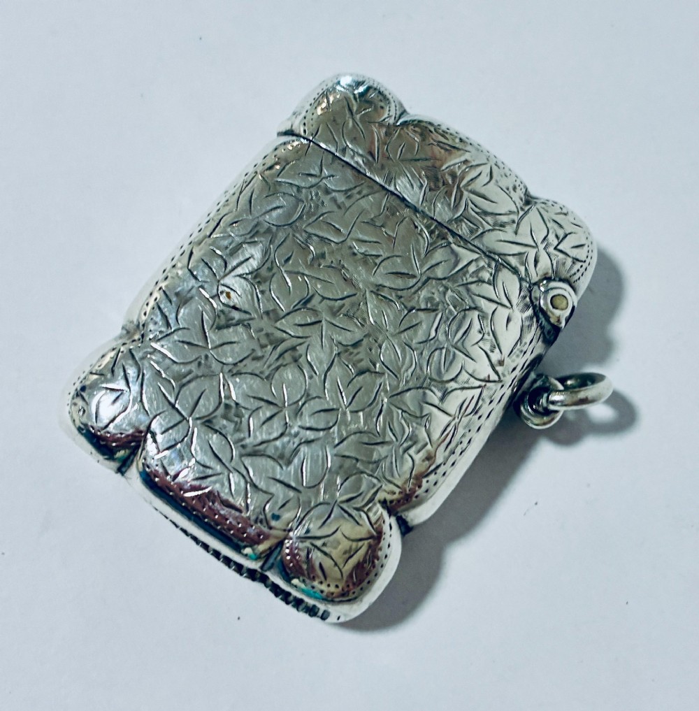 A Victorian sterling silver vesta case featuring hand-engraved foliage motifs, a central shield cartouche, lobed edges, and a side attachment ring. View V