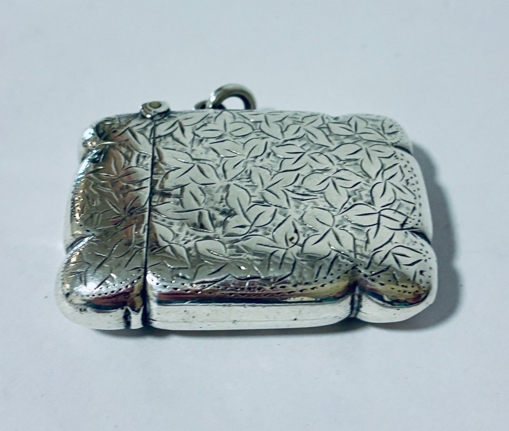 A Victorian sterling silver vesta case featuring hand-engraved foliage motifs, a central shield cartouche, lobed edges, and a side attachment ring. View VI