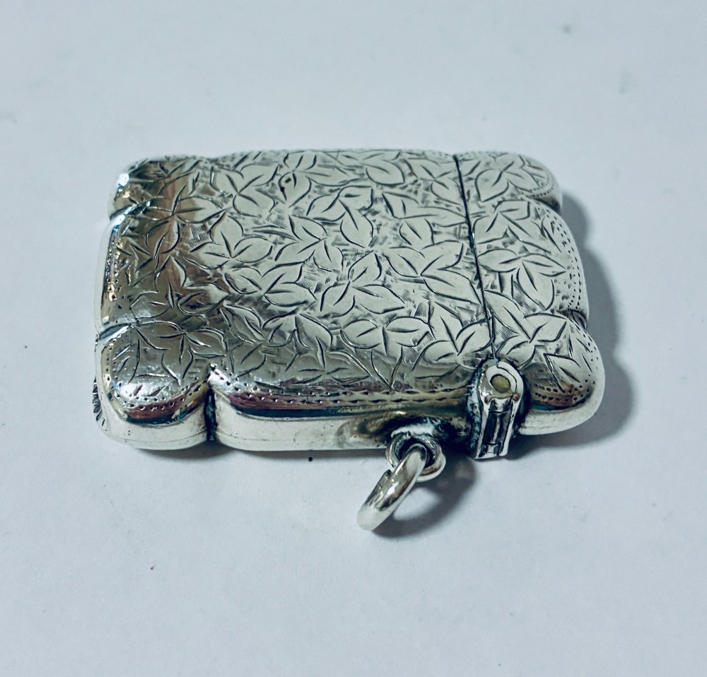 A Victorian sterling silver vesta case featuring hand-engraved foliage motifs, a central shield cartouche, lobed edges, and a side attachment ring. View VII