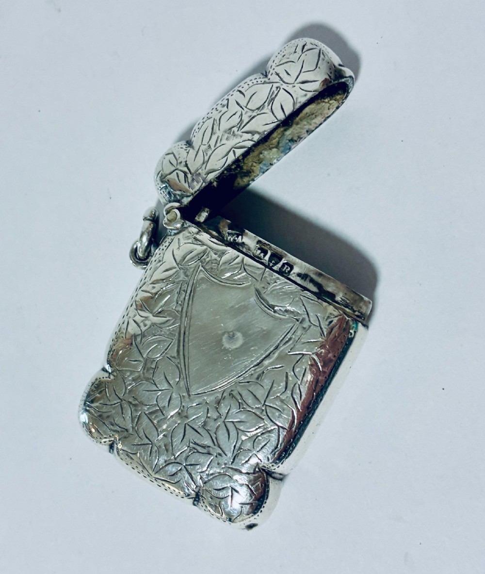 A Victorian sterling silver vesta case featuring hand-engraved foliage motifs, a central shield cartouche, lobed edges, and a side attachment ring. View VIII