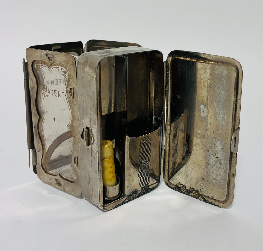 An open nickel-plated folding pocket lantern containing a small wax candle and a glass panel stamped with patent text.