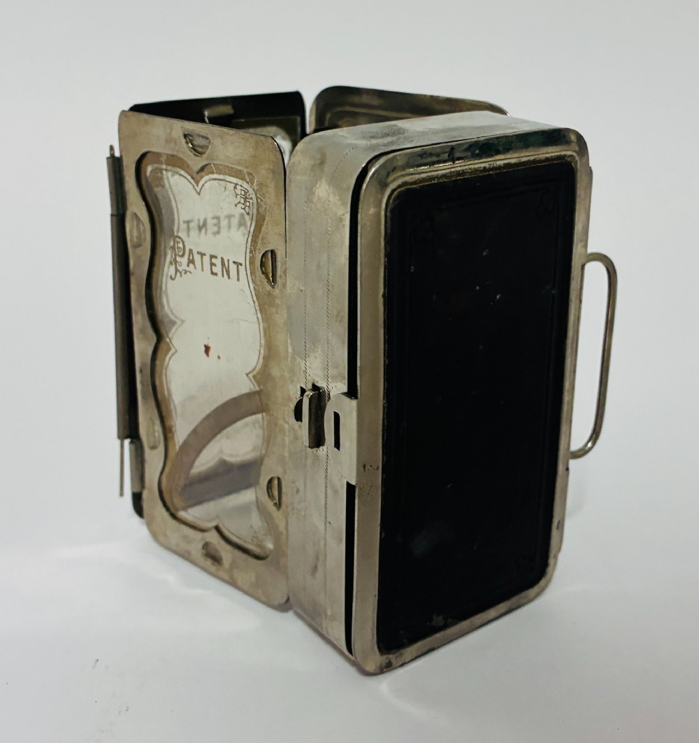 An open nickel-plated folding pocket lantern containing a small wax candle and a glass panel stamped with patent text. View III