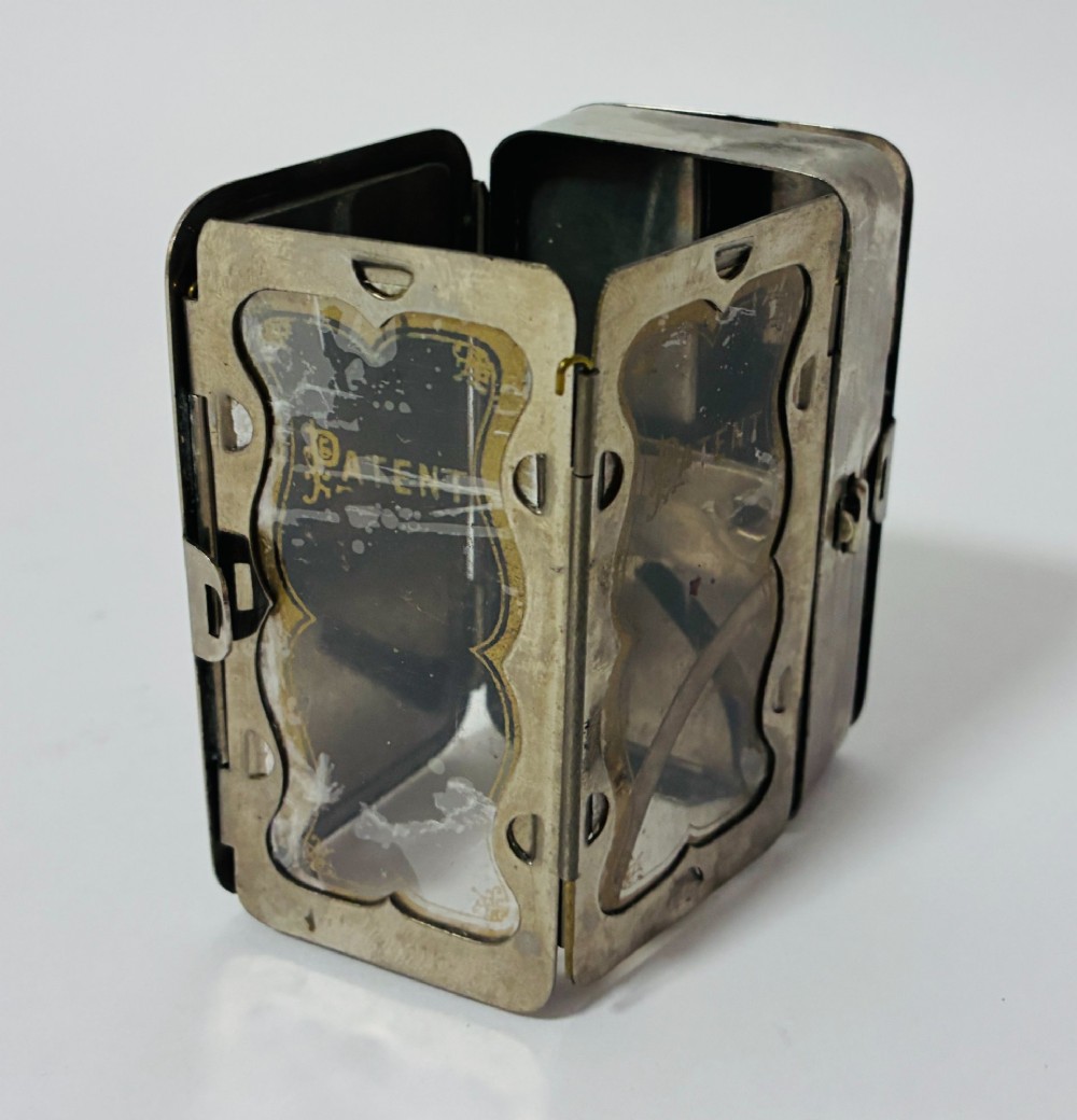 An open nickel-plated folding pocket lantern containing a small wax candle and a glass panel stamped with patent text. View IV