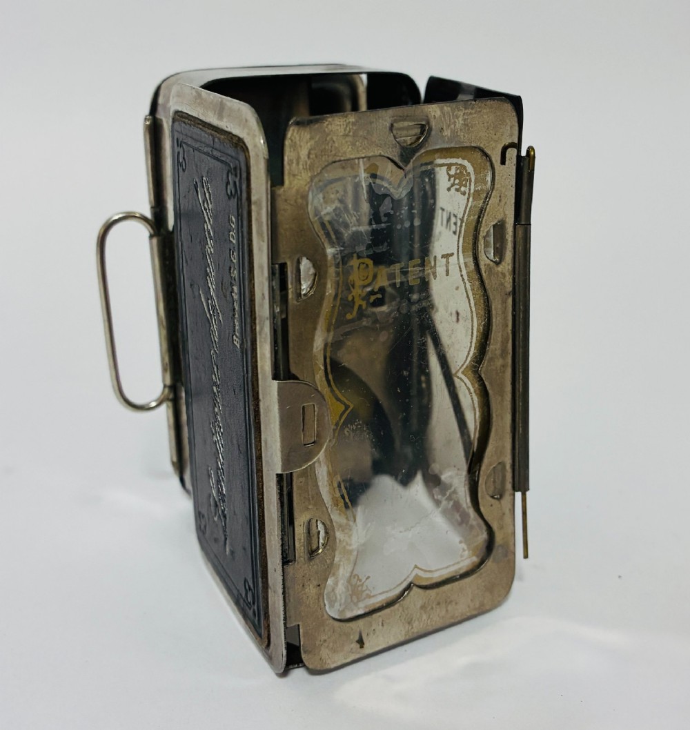 An open nickel-plated folding pocket lantern containing a small wax candle and a glass panel stamped with patent text. View V