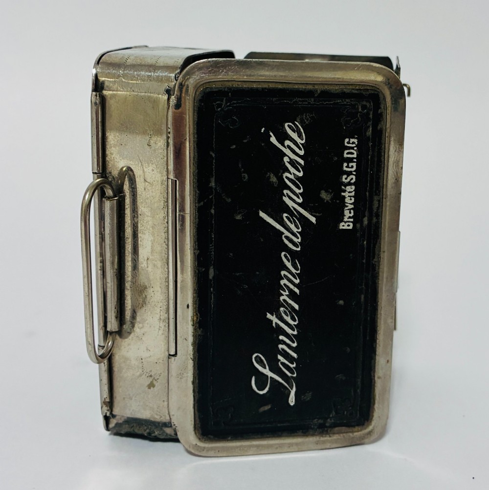 An open nickel-plated folding pocket lantern containing a small wax candle and a glass panel stamped with patent text. View VI
