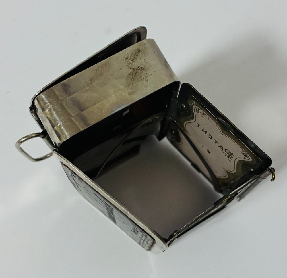 An open nickel-plated folding pocket lantern containing a small wax candle and a glass panel stamped with patent text. View VII