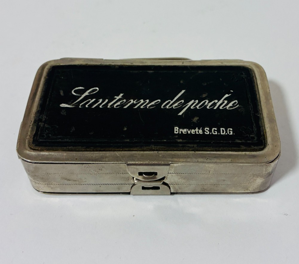 An open nickel-plated folding pocket lantern containing a small wax candle and a glass panel stamped with patent text. View VIII