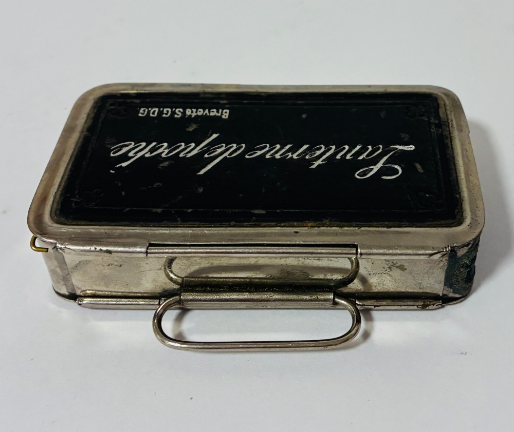 An open nickel-plated folding pocket lantern containing a small wax candle and a glass panel stamped with patent text. View IX