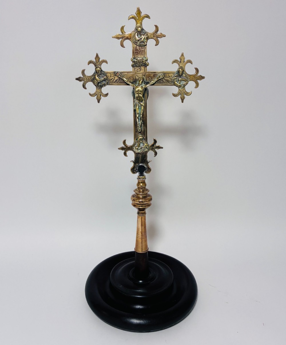 An antique brass processional cross featuring a detailed crucifix figure, fleur-de-lis terminal accents, and engraved motifs, mounted on a turned dark wood stand.