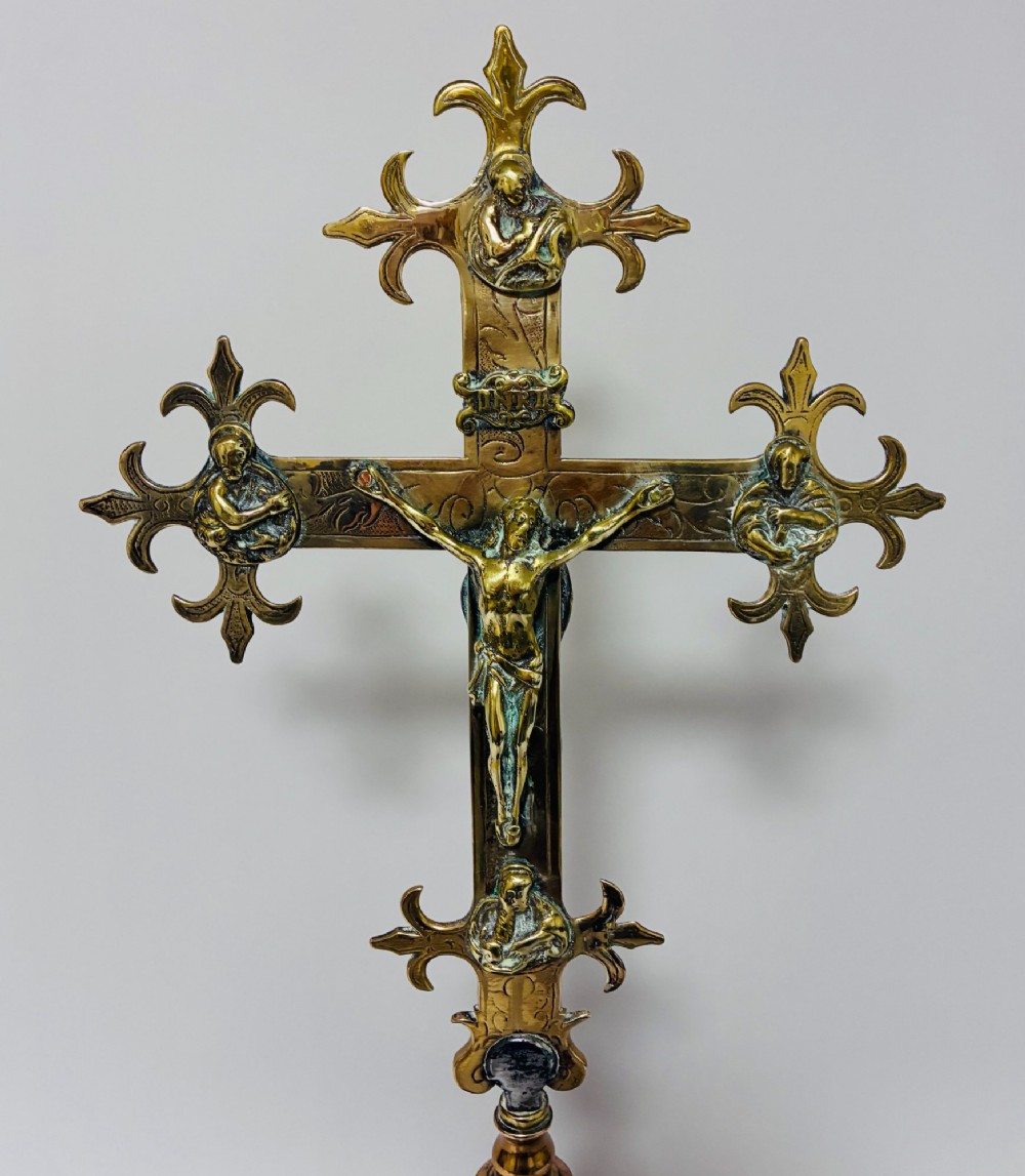 An antique brass processional cross featuring a detailed crucifix figure, fleur-de-lis terminal accents, and engraved motifs, mounted on a turned dark wood stand. View II