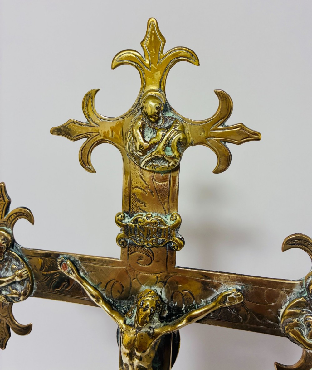 An antique brass processional cross featuring a detailed crucifix figure, fleur-de-lis terminal accents, and engraved motifs, mounted on a turned dark wood stand. View IV