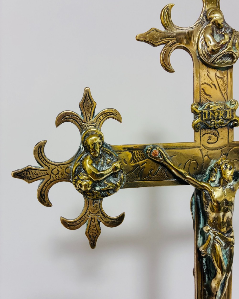 An antique brass processional cross featuring a detailed crucifix figure, fleur-de-lis terminal accents, and engraved motifs, mounted on a turned dark wood stand. View V