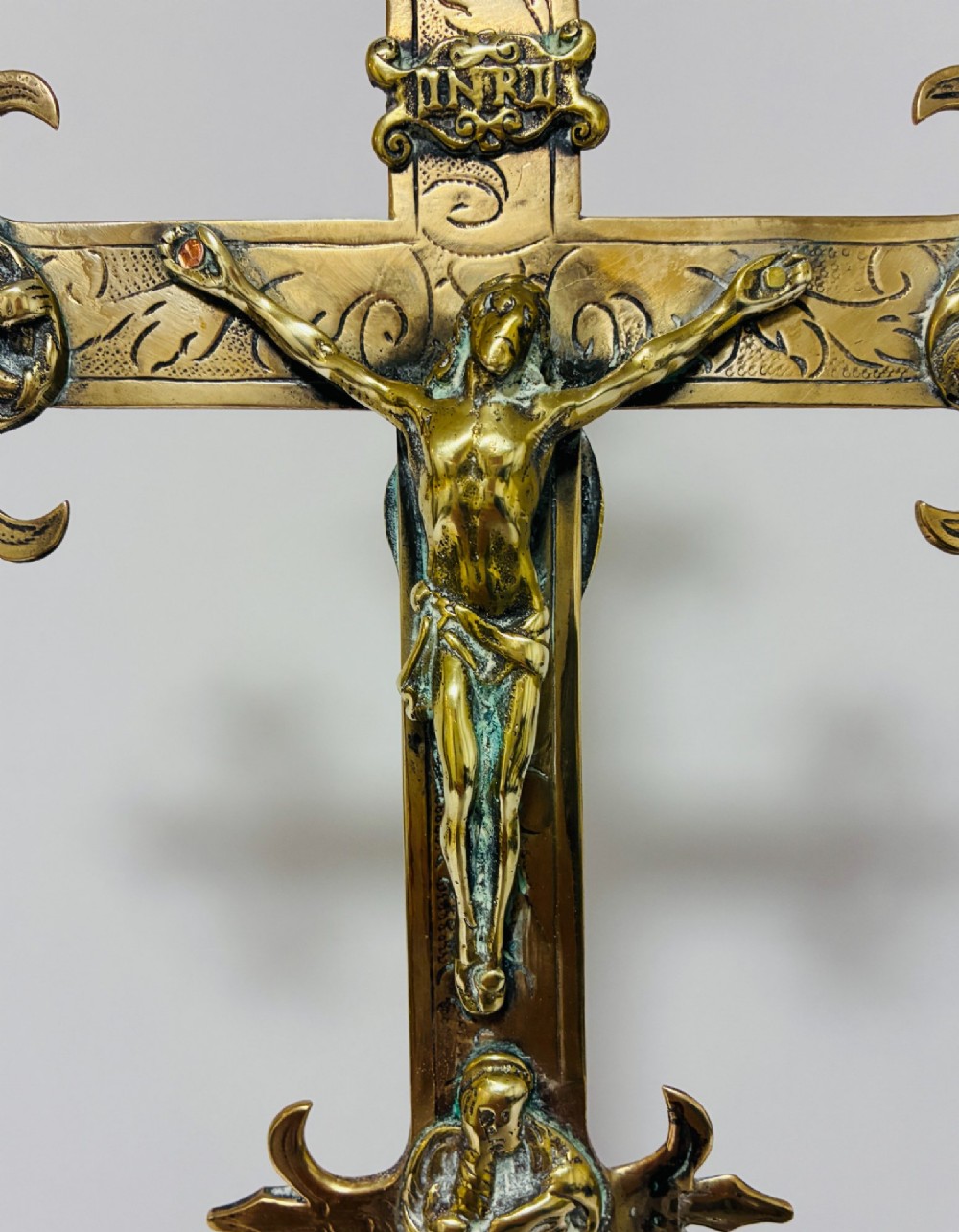 An antique brass processional cross featuring a detailed crucifix figure, fleur-de-lis terminal accents, and engraved motifs, mounted on a turned dark wood stand. View VI