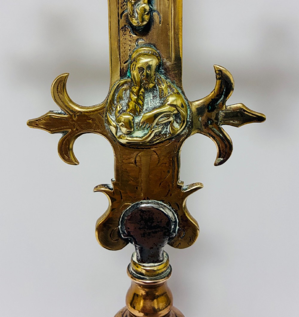 An antique brass processional cross featuring a detailed crucifix figure, fleur-de-lis terminal accents, and engraved motifs, mounted on a turned dark wood stand. View VII