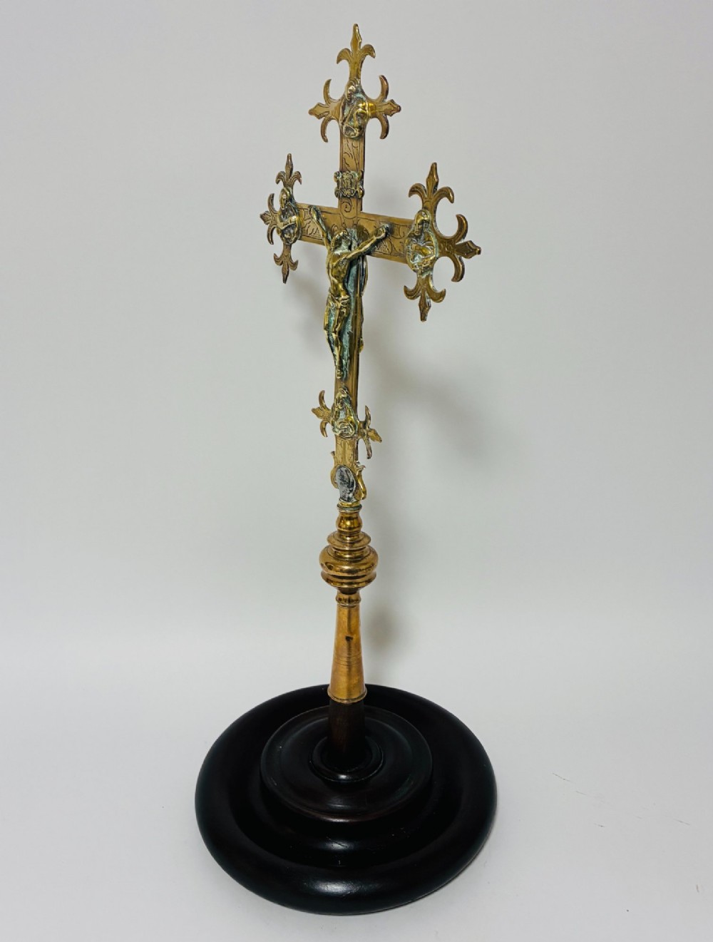 An antique brass processional cross featuring a detailed crucifix figure, fleur-de-lis terminal accents, and engraved motifs, mounted on a turned dark wood stand. View X