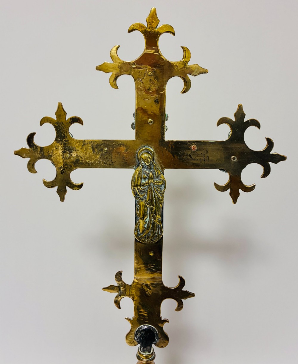 An antique brass processional cross featuring a detailed crucifix figure, fleur-de-lis terminal accents, and engraved motifs, mounted on a turned dark wood stand. View XII