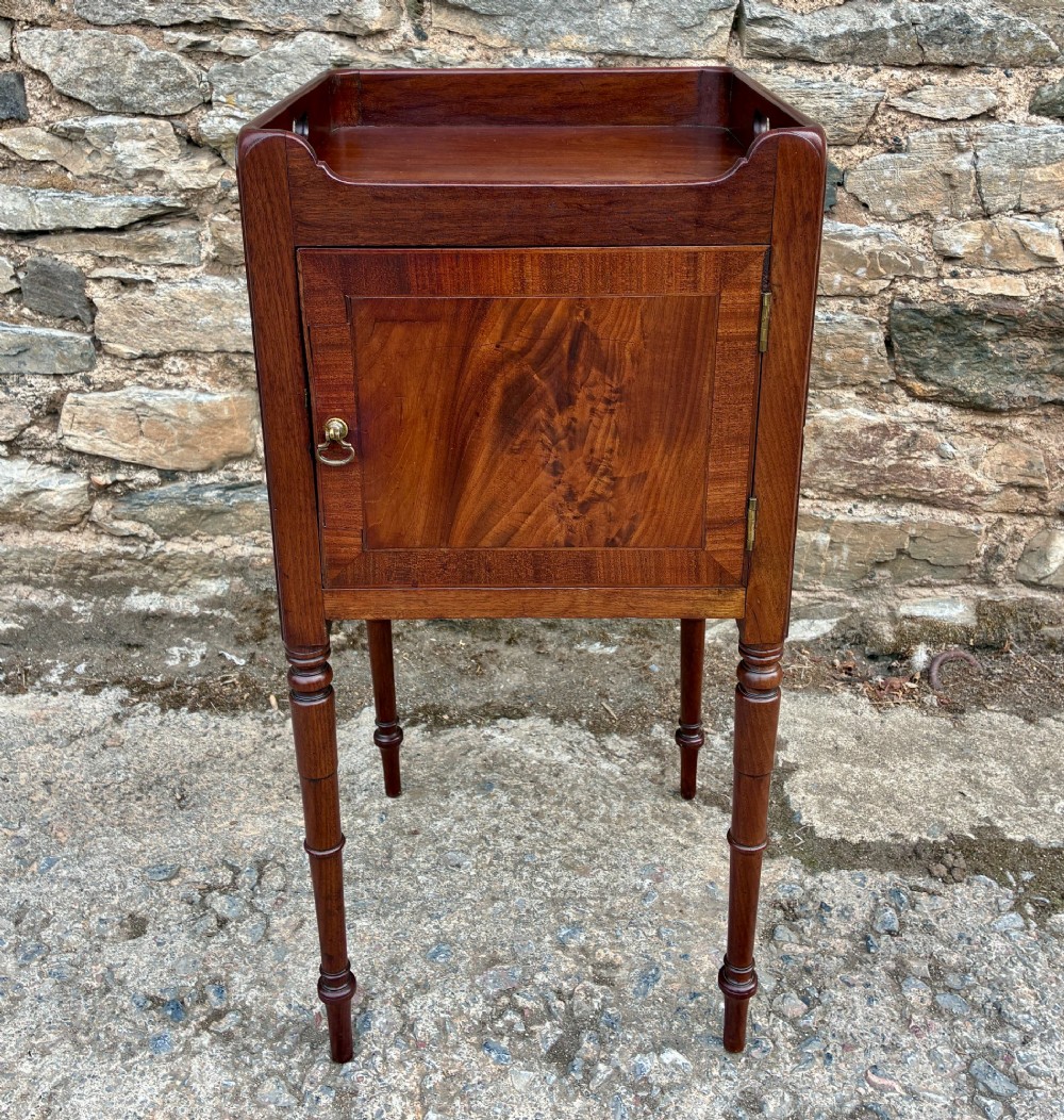 A Georgian mahogany pot cupboard featuring a tray top gallery with cut-out handles, a single veneered door, and turned legs.
