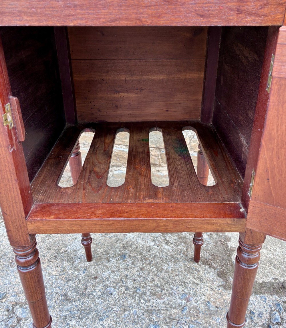 A Georgian mahogany pot cupboard featuring a tray top gallery with cut-out handles, a single veneered door, and turned legs. View V