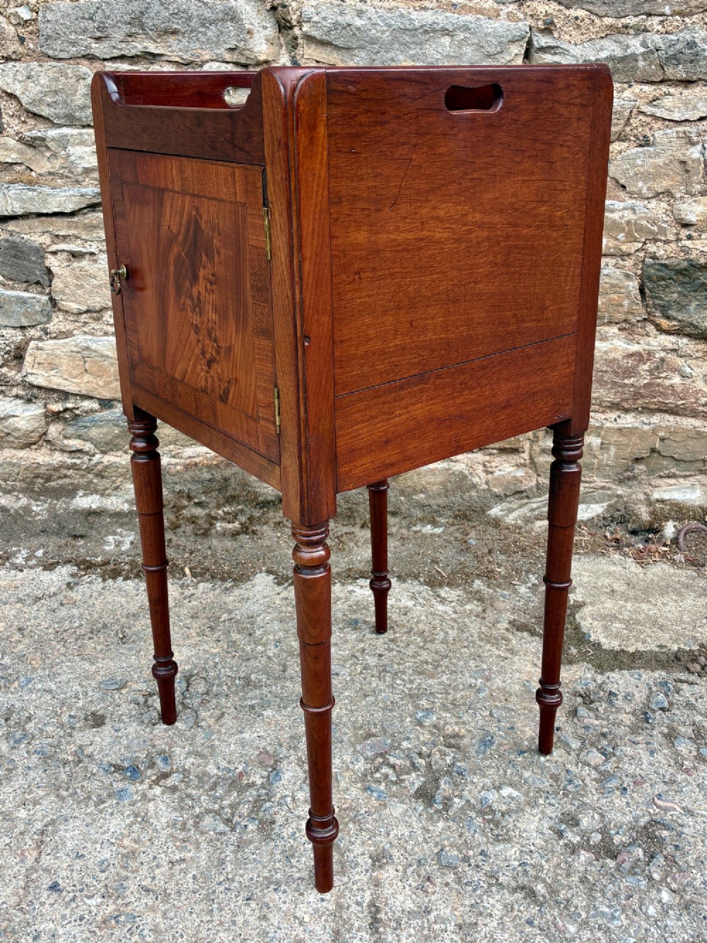 A Georgian mahogany pot cupboard featuring a tray top gallery with cut-out handles, a single veneered door, and turned legs. View VI