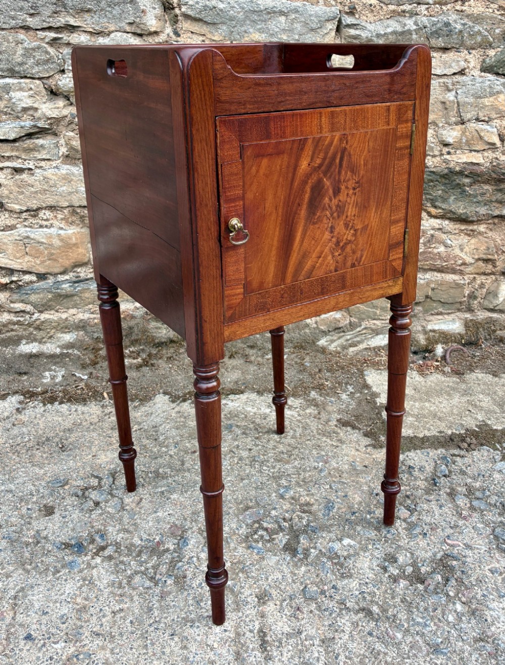 A Georgian mahogany pot cupboard featuring a tray top gallery with cut-out handles, a single veneered door, and turned legs. View X