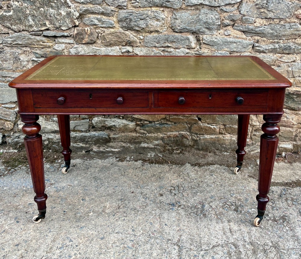 A Victorian mahogany writing table featuring a green gilt-tooled leather writing surface, two drawers with turned knob handles, and turned tapered legs resting on ceramic castors.