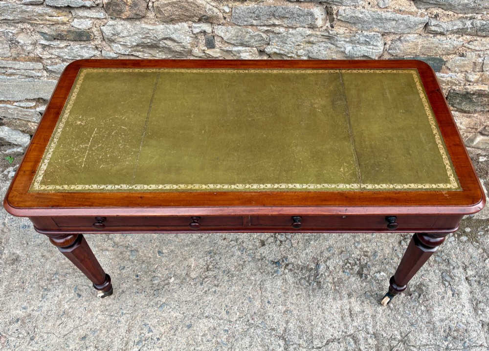 A Victorian mahogany writing table featuring a green gilt-tooled leather writing surface, two drawers with turned knob handles, and turned tapered legs resting on ceramic castors. View II