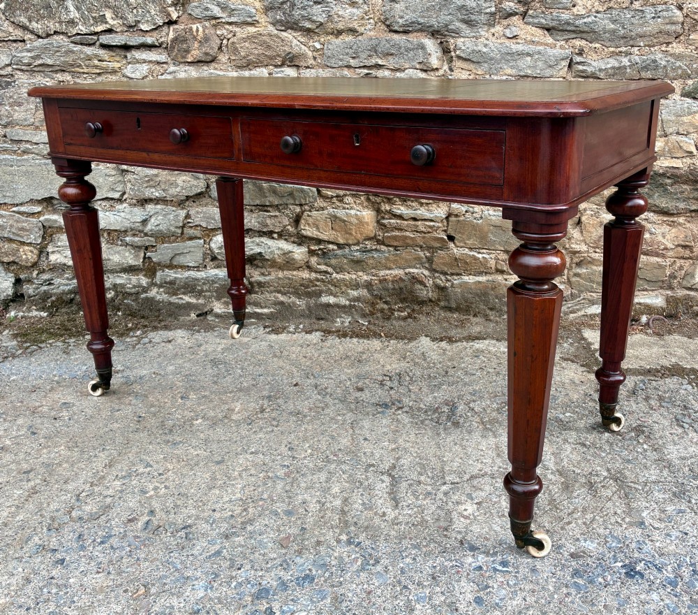 A Victorian mahogany writing table featuring a green gilt-tooled leather writing surface, two drawers with turned knob handles, and turned tapered legs resting on ceramic castors. View VI