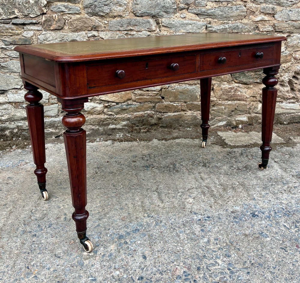 A Victorian mahogany writing table featuring a green gilt-tooled leather writing surface, two drawers with turned knob handles, and turned tapered legs resting on ceramic castors. View XII