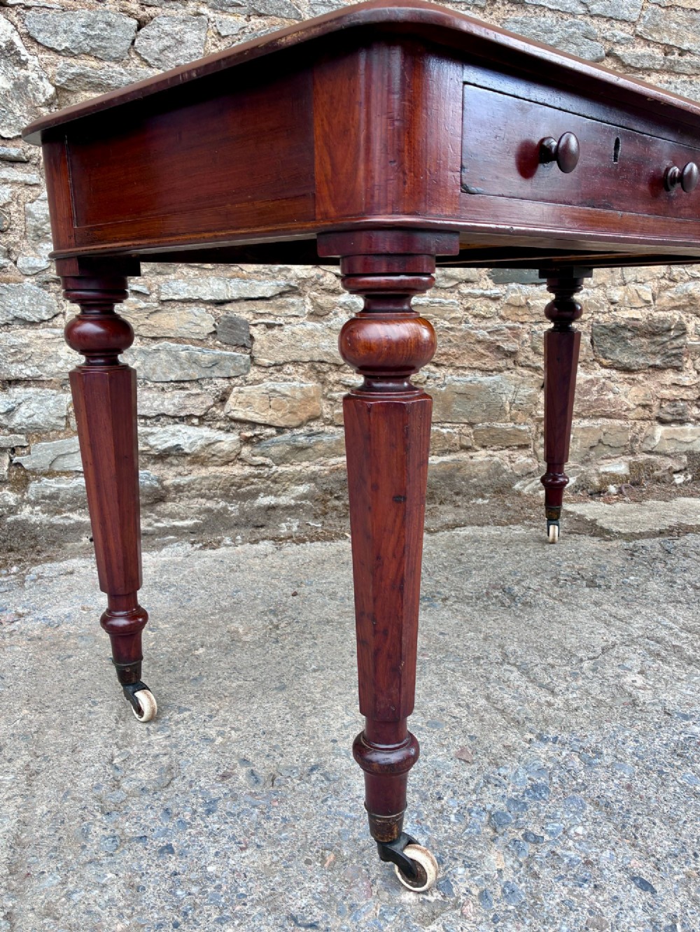 A Victorian mahogany writing table featuring a green gilt-tooled leather writing surface, two drawers with turned knob handles, and turned tapered legs resting on ceramic castors. View XIII