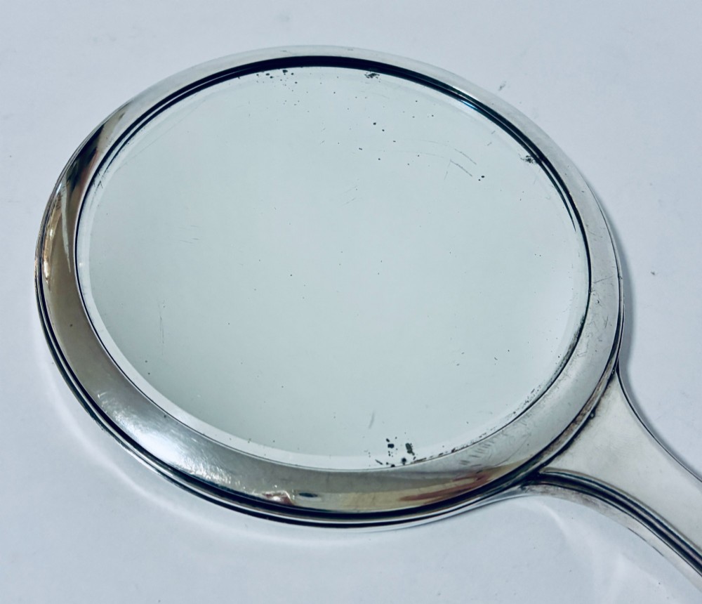 An antique oval silver hand mirror resting horizontally against a plain background. View IV