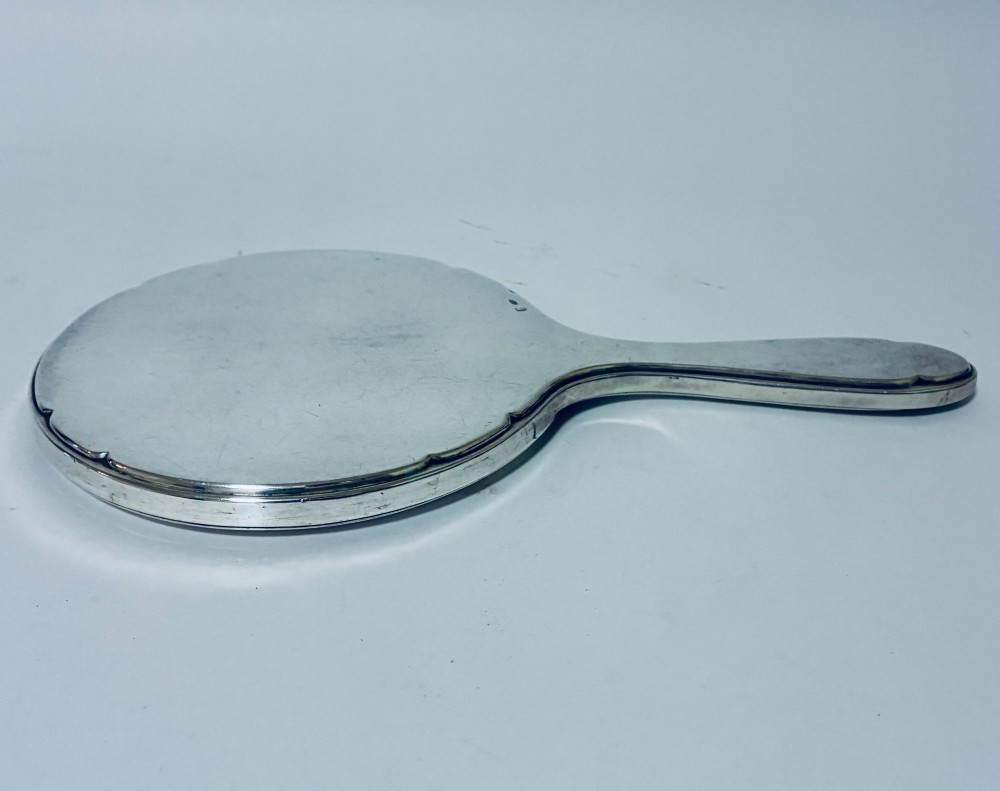 An antique oval silver hand mirror resting horizontally against a plain background. View V