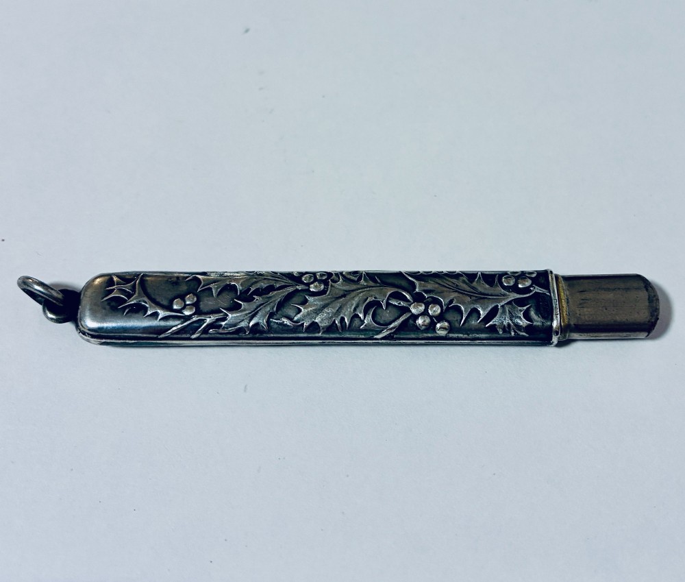 An antique silver cylindrical pencil holder featuring repousse holly leaf and berry decoration with a suspension ring attached to the end.