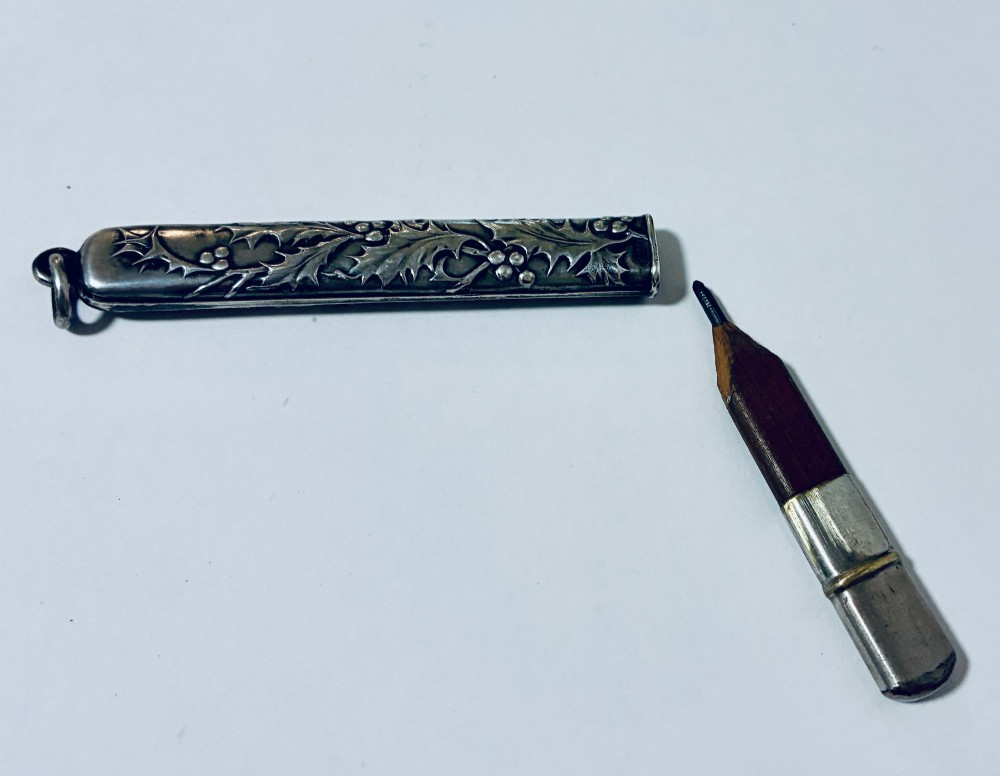 An antique silver cylindrical pencil holder featuring repousse holly leaf and berry decoration with a suspension ring attached to the end. View II