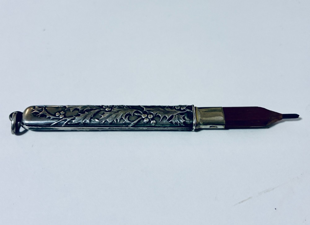 An antique silver cylindrical pencil holder featuring repousse holly leaf and berry decoration with a suspension ring attached to the end. View III