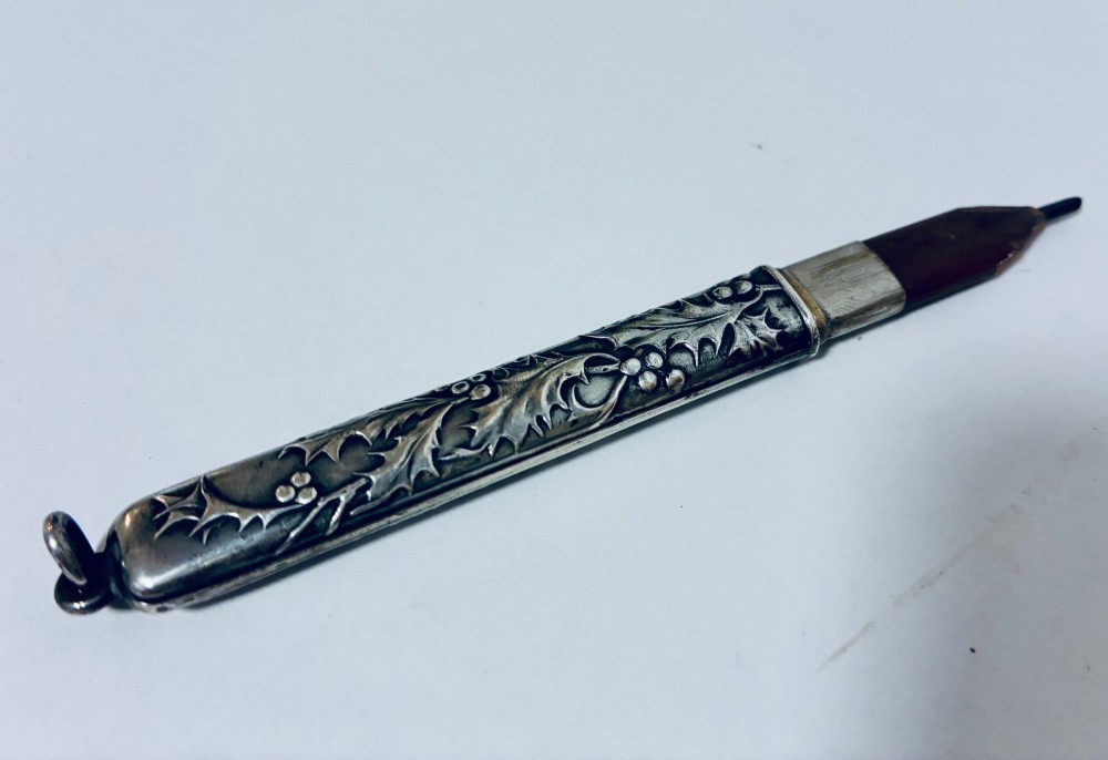 An antique silver cylindrical pencil holder featuring repousse holly leaf and berry decoration with a suspension ring attached to the end. View IV