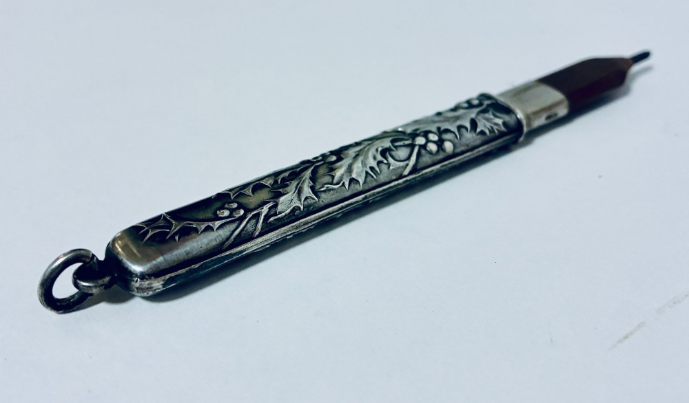 An antique silver cylindrical pencil holder featuring repousse holly leaf and berry decoration with a suspension ring attached to the end. View V