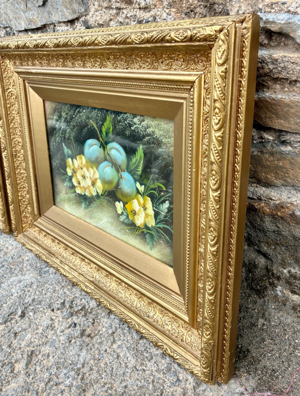 A matching pair of Victorian still life oil paintings depicting plums, primroses, and a bird nest in ornate gold gilded frames. View X