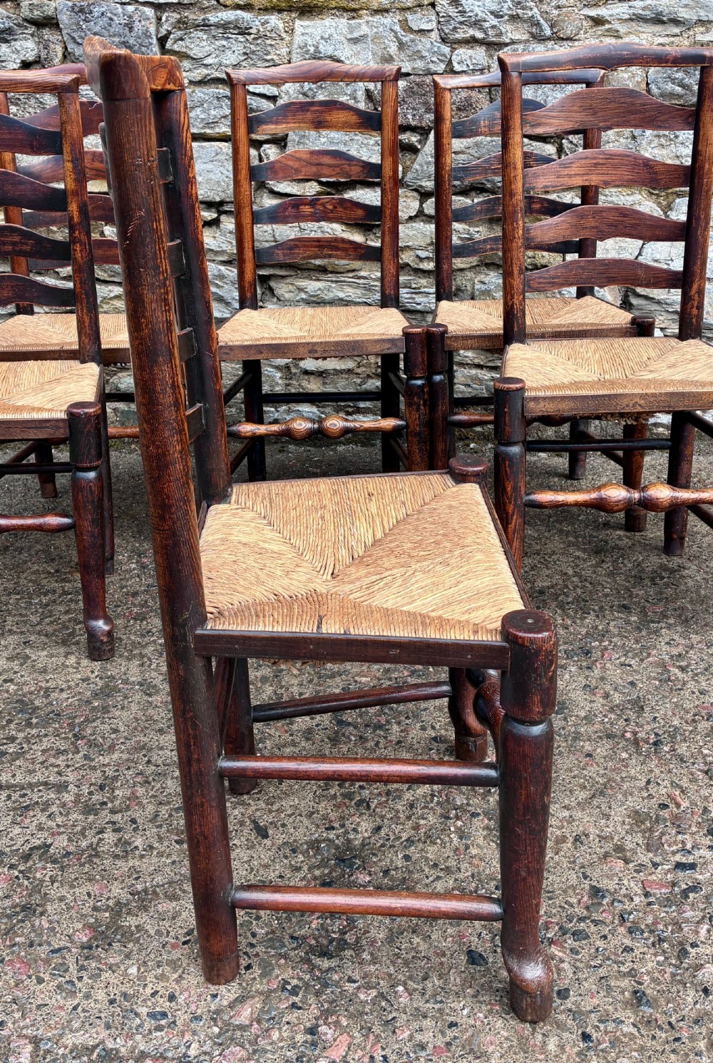 A set of eight 19th-century dark timber Lancashire ladderback dining chairs featuring wavy back slats, turned front stretchers, and woven rush seats, comprising two carver armchairs and six side chairs. View XII