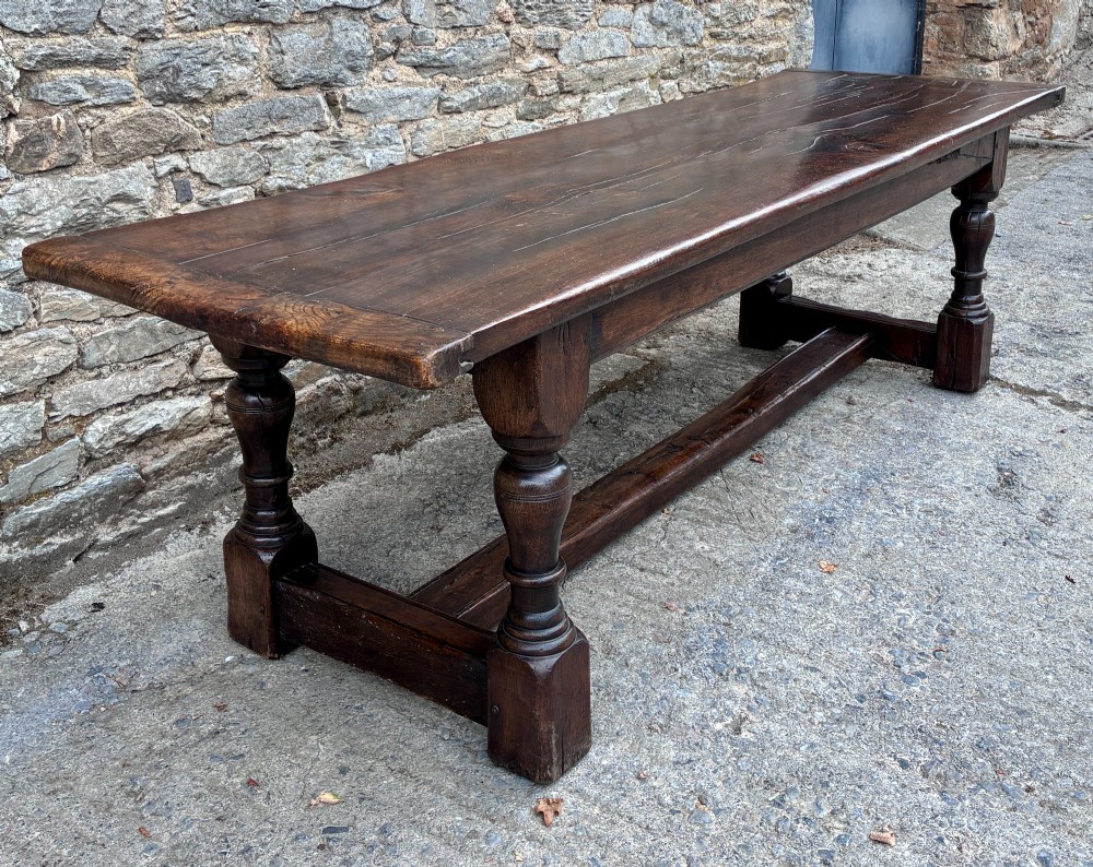 A long dark oak refectory dining table featuring a solid plank top, cleated ends, turned legs, and a low stretcher rail. View III