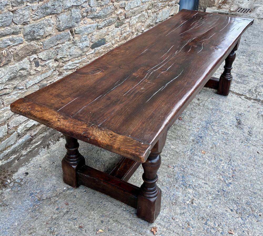 A long dark oak refectory dining table featuring a solid plank top, cleated ends, turned legs, and a low stretcher rail. View IV