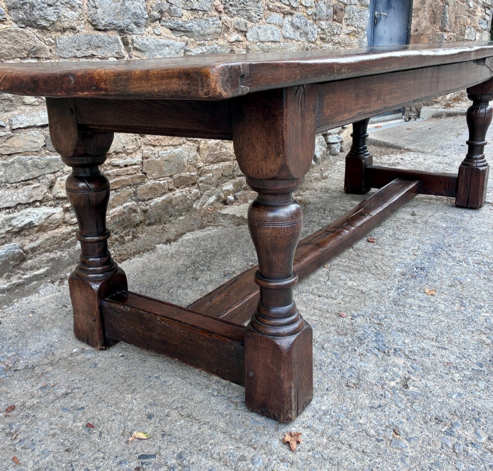 A long dark oak refectory dining table featuring a solid plank top, cleated ends, turned legs, and a low stretcher rail. View V